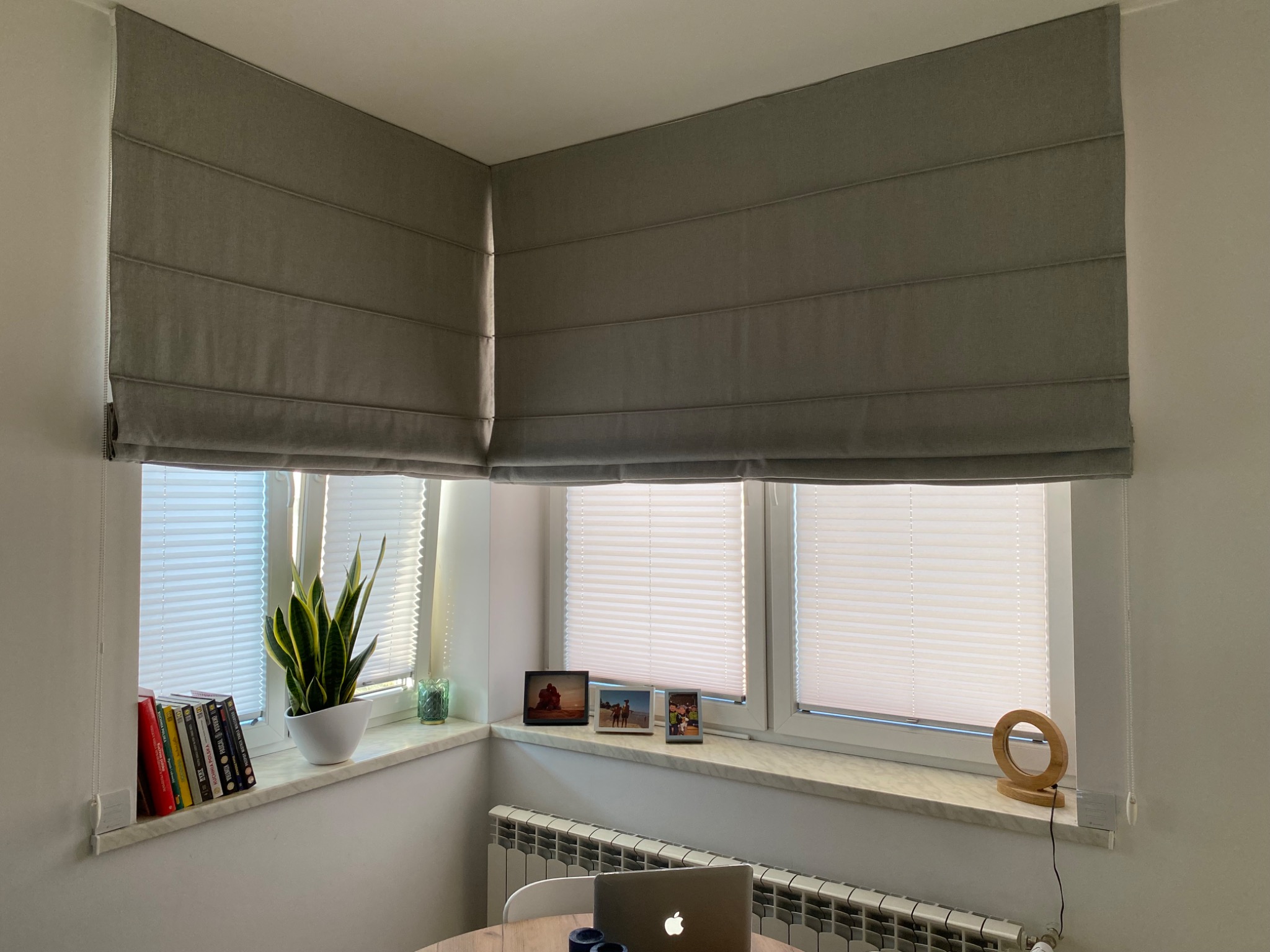 Szare, materiałowe zasłony w stylu rzymskim zamontowane w rogu pokoju, obok okna z plisami, na parapecie książki, roślina i ramki ze zdjęciami.
