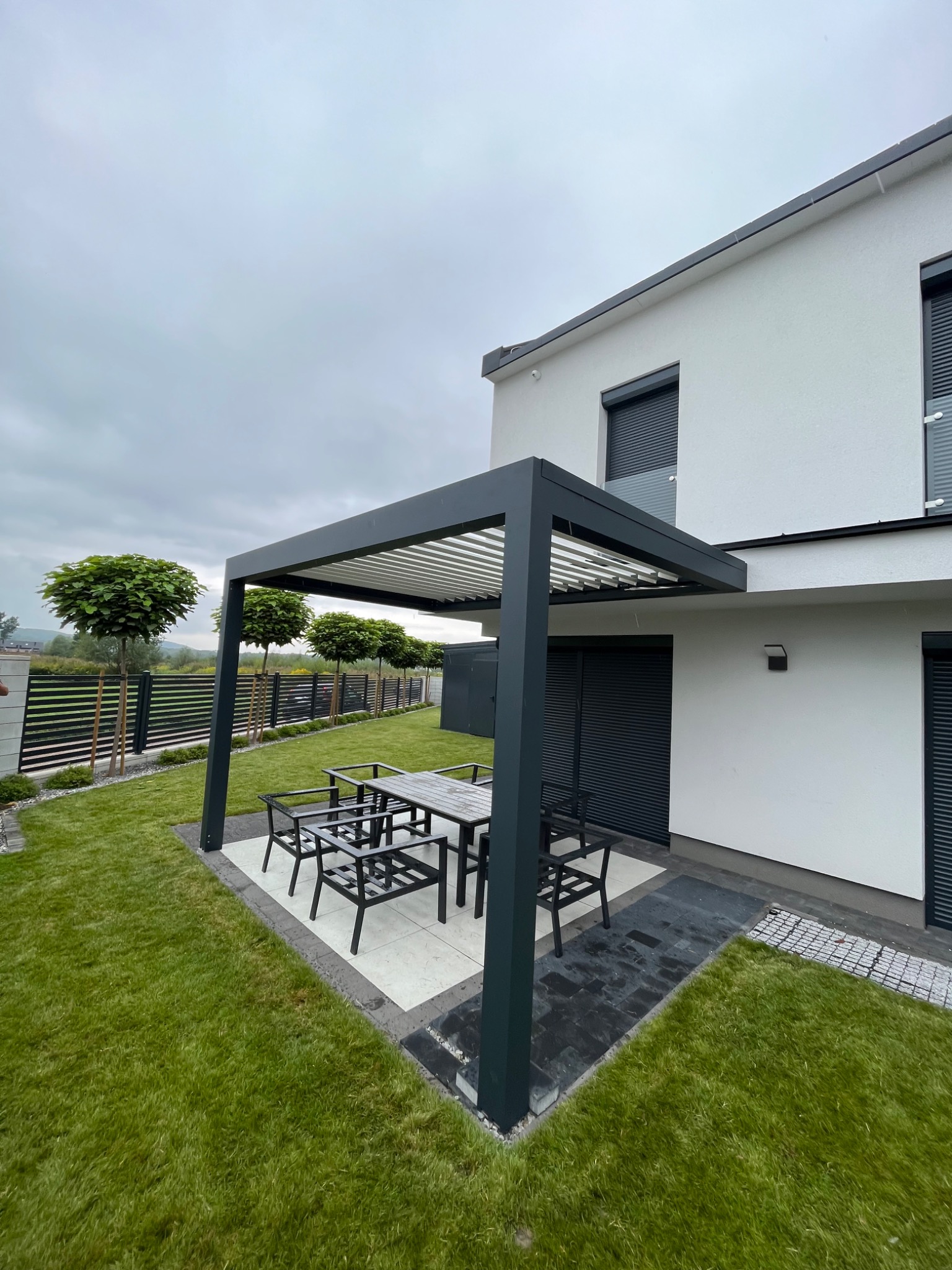 Nowoczesny dom z szarymi ramami okiennymi i zewnętrznymi osłonami, pergola z miejscem do siedzenia na zewnątrz, zadbany trawnik i ozdobne drzewa w rzędzie.