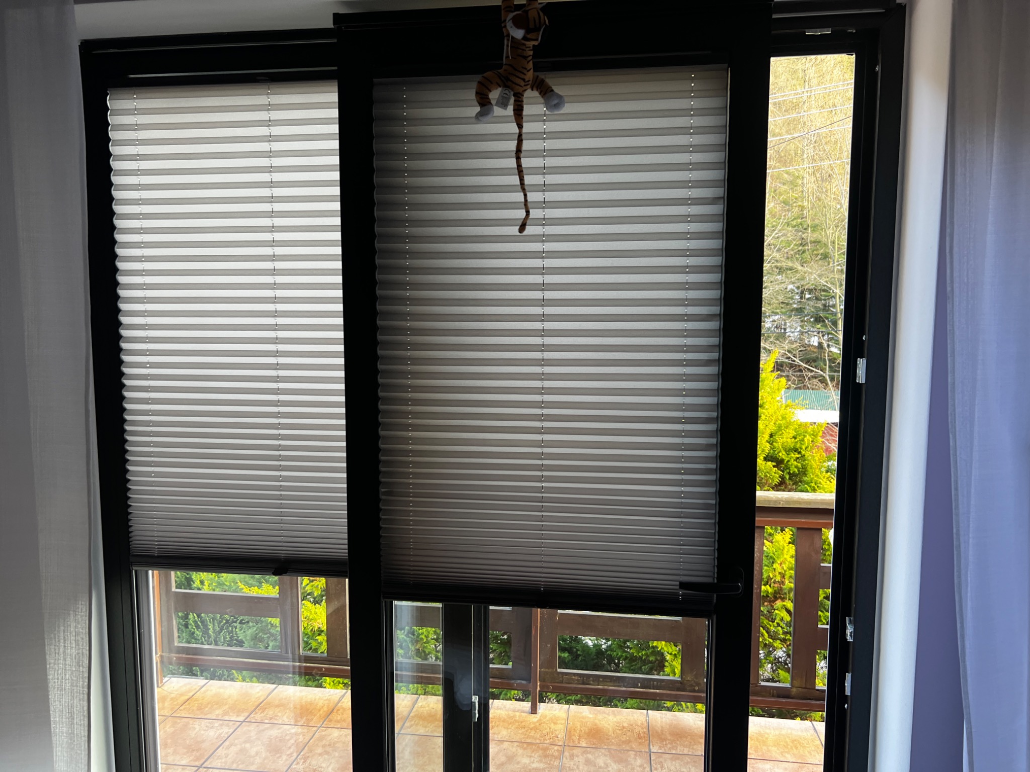 Zamknięte okno balkonowe z szarymi plisami, na jednej z nich zawieszona pluszowa zabawka tygryska, widok na balkon i zieleń za oknem.