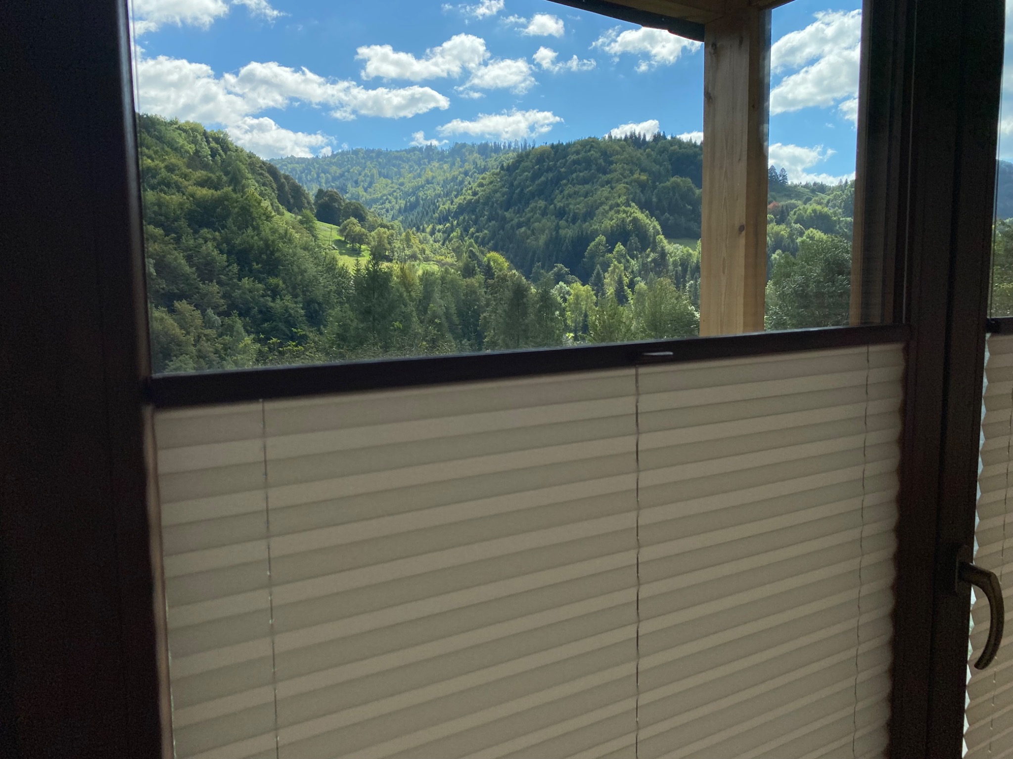 Widok z okna z założonymi jasnymi plisami na zalesione wzgórza i błękitne niebo z chmurami.