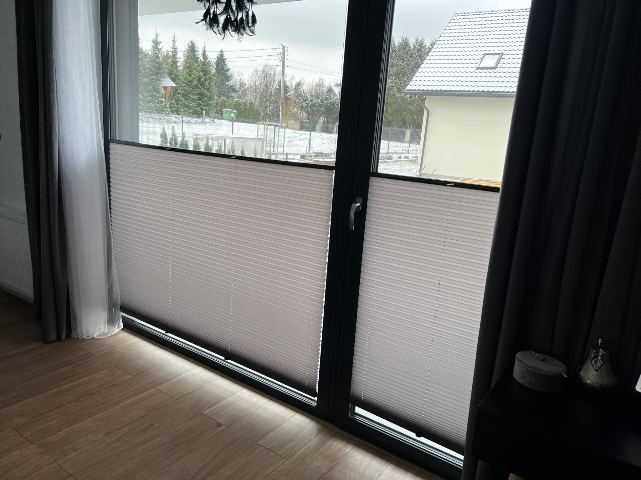 Jasne plisy okienne w nowoczesnym salonie z widokiem na zaśnieżony ogród i dom.