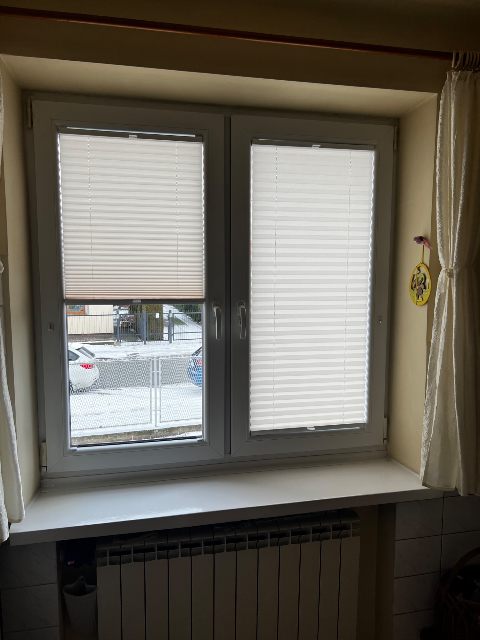 Okno z zamontowanymi białymi plisami, jedna z nich częściowo opuszczona, widok na zasnieżony krajobraz za oknem.