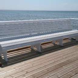 Biała ławka z drewnianych desek, umieszczona na drewnianym molo z widokiem na spokojne morze w słoneczny dzień.