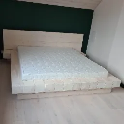 Drewniane łóżko z jasnych belek, z materacem, ustawione na podłodze z desek w pokoju z ciemnozieloną i białą ścianą.