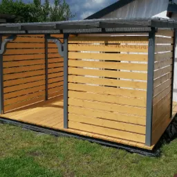 Nowoczesna, drewniana konstrukcja z poziomymi panelami, szarymi ramami i przezroczystym zadaszeniem, stojąca na trawniku obok domu.