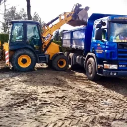 Żółto-czarna koparko-ładowarka JCB załadowuje niebieską wywrotkę Scania na placu budowy, widoczne ślady opon w ziemi.