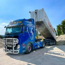 Niebieska ciężarówka Volvo z naczepą wywrotką, z logo firmy DOMIN, wysypuje piasek na betonowy plac, widoczne zabezpieczenie przednie z białej kraty, błękitne niebo w tle.