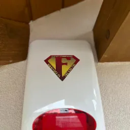 Biała obudowa sygnalizatora alarmowego z czerwonym światłem, zamontowana w rogu pomieszczenia pod drewnianą belką, z naklejoną naklejką firmy Systemy Zabezpieczeń FOXX.