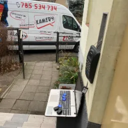 Montaż domofonu cyfrowego przy wejściu do budynku, z laptopem i kluczami na schodach, na tle firmowego samochodu z reklamą usług.