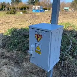 Szara skrzynka elektryczna zamontowana na metalowym słupku na trawiastym terenie, z kluczem w zamku i tabliczką ostrzegawczą o wysokim napięciu.