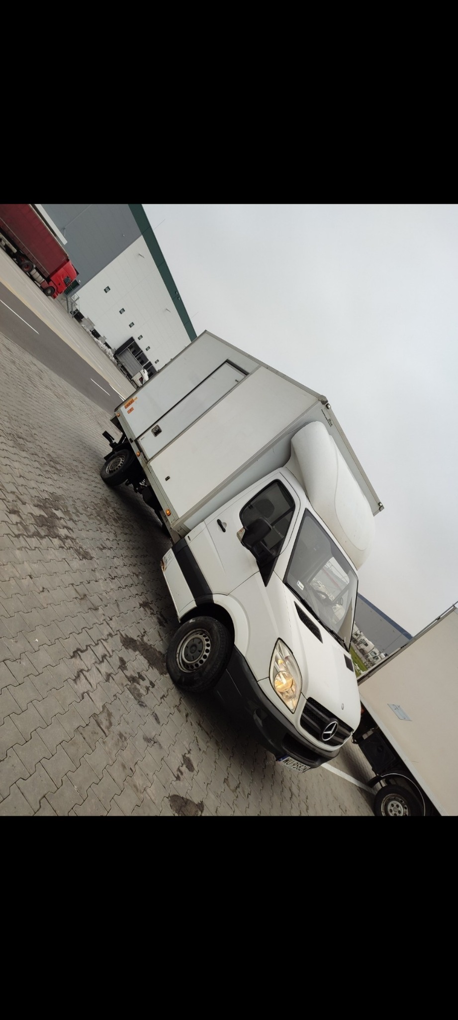 Biały samochód dostawczy Mercedes-Benz z zabudową kontenerową na parkingu z kostki brukowej, widoczne inne pojazdy i hala magazynowa w tle.
