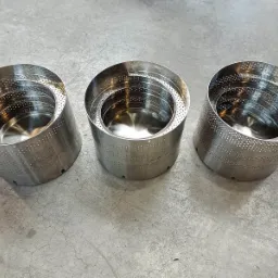 Trzy identyczne, metalowe, perforowane cylindry ustawione obok siebie na betonowej posadzce. Wnętrze każdego cylindra jest gładkie i błyszczące, a perforacja tworzy regularny wzór.