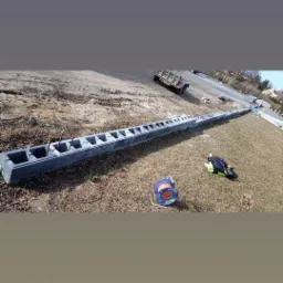 Długa, szara, betonowa forma fundamentowa z otworami ustawiona na ziemi, obok leży zwinięty kabel i torba robocza, w tle przyczepa i domy.