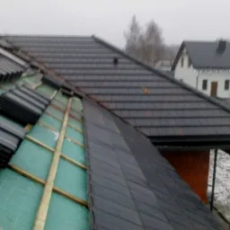 Częściowo pokryty dach z czarną dachówką, widoczne drewniane łaty i zielona membrana dachowa, w tle dom z ciemnym dachem i białą elewacją w pochmurny dzień.