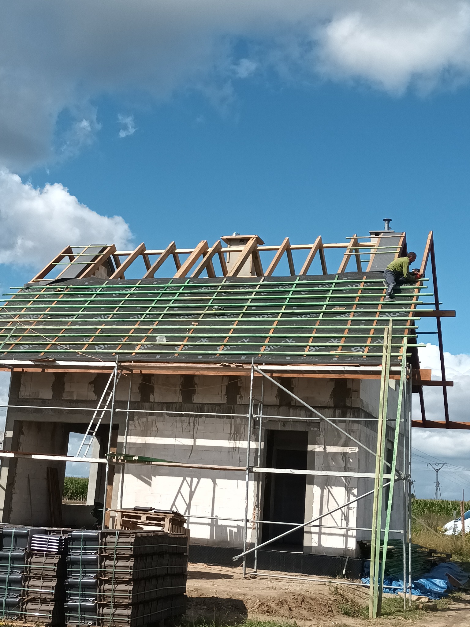 Dekarz na dachu w trakcie krycia, widoczne łaty, membrana dachowa, komin, palety z dachówką na ziemi, rusztowanie i fragmenty niezakończonej elewacji budynku.