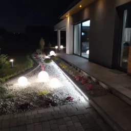Nocne ujęcie posesji z oświetleniem ogrodowym: kuliste lampy na żwirowym podłożu, listwa LED wzdłuż rabaty kwiatowej i punktowe oświetlenie elewacji budynku.