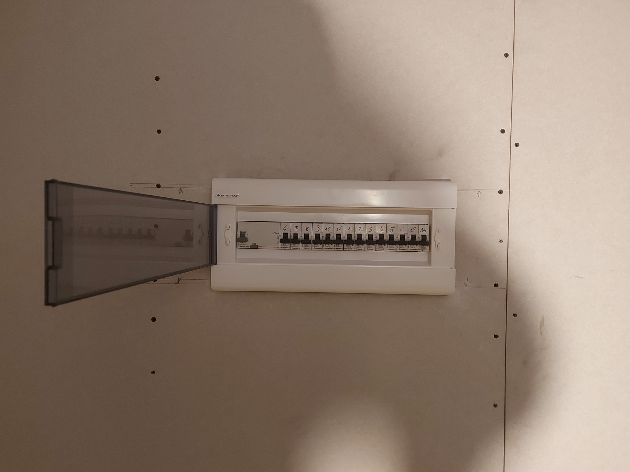 Rozdzielnica elektryczna z otwartą osłoną na ścianie z płyt gipsowo-kartonowych, widoczne oznaczenia obwodów i ślady po montażu.