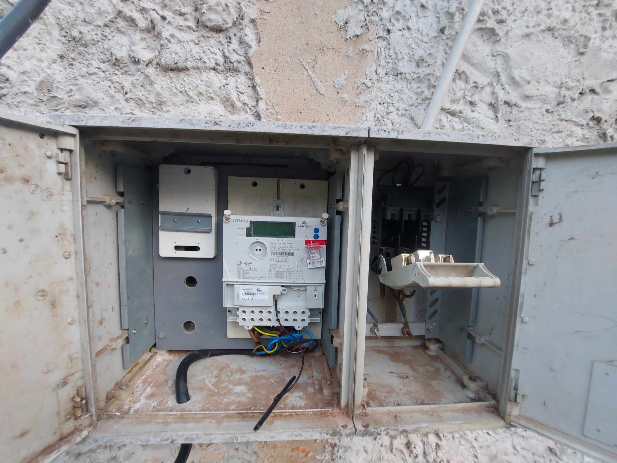 Otwarta skrzynka elektryczna z licznikiem OTUS 3 na tle surowej, nierównej ściany z widocznymi przewodami i elementami instalacji.