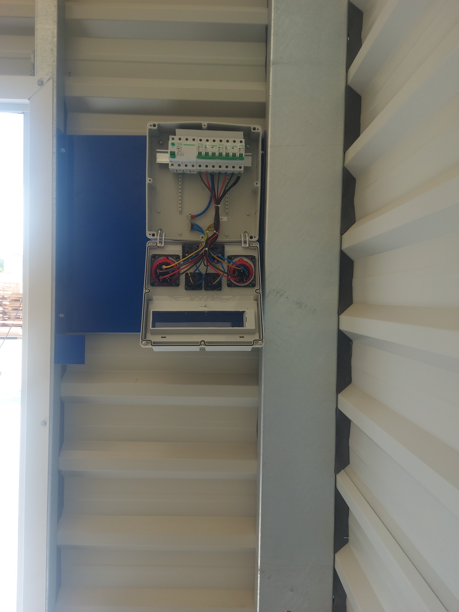 Otwarta skrzynka elektryczna z widocznymi przewodami i bezpiecznikami, zamontowana na konstrukcji z blachy trapezowej w jasnym pomieszczeniu.