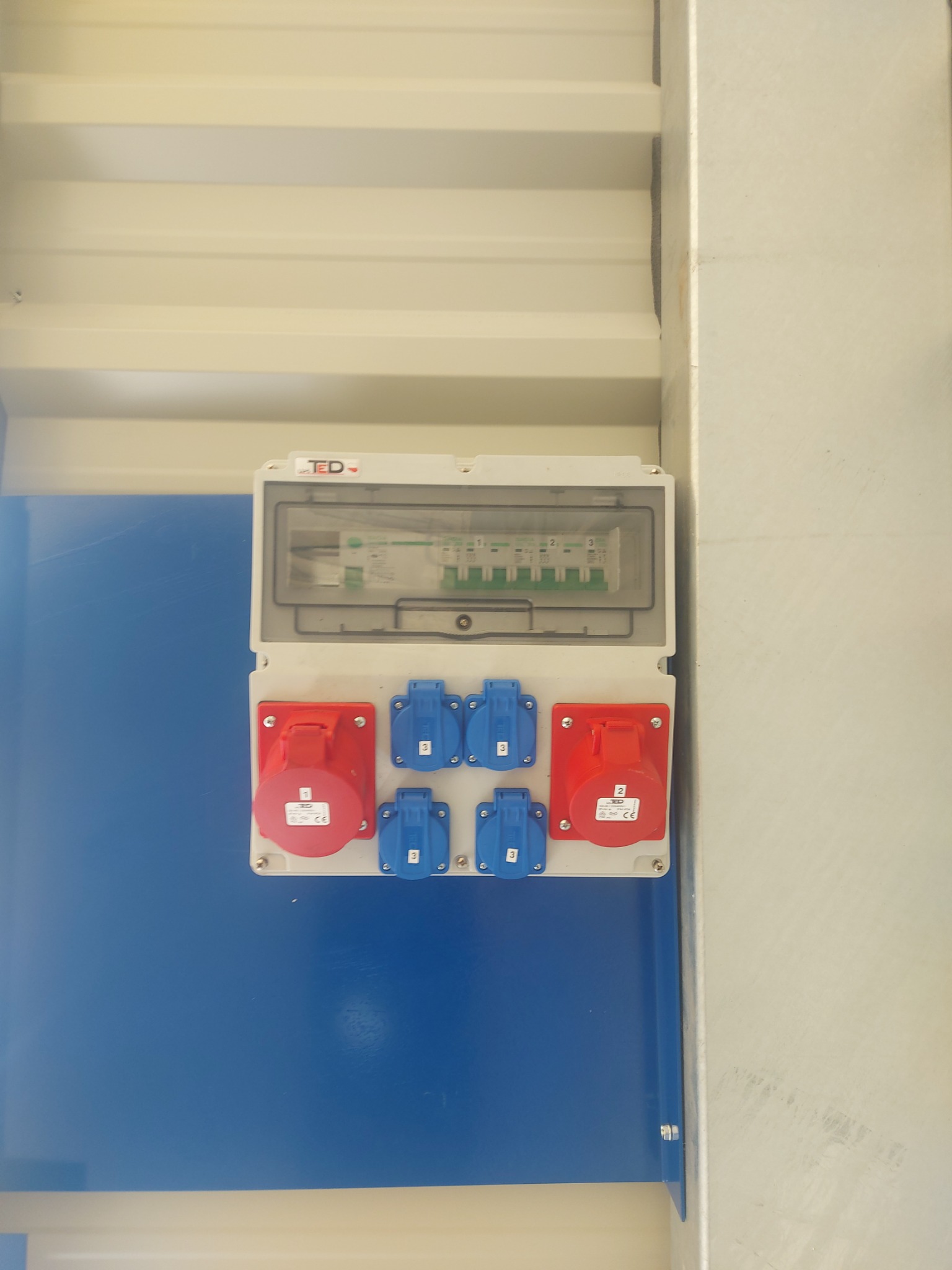 Rozdzielnica elektryczna z bezpiecznikami i gniazdami siłowymi (czerwone i niebieskie) zamontowana na niebieskiej ścianie wewnątrz kontenera.
