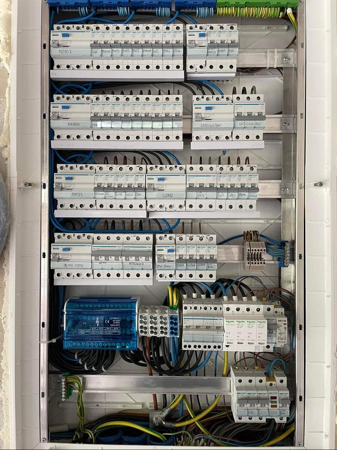 Rozdzielnica elektryczna z zainstalowanymi bezpiecznikami i okablowaniem, etykietami opisującymi obwody (kuchnia, parter, garaż), widoczne elementy zabezpieczeń przepięciowych i listwy zaciskowe.