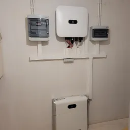 Komponenty instalacji fotowoltaicznej na białej ścianie: inwerter Huawei, rozdzielnice elektryczne i akumulator energii, połączone białymi rurami i listwami.