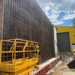 Siatka zbrojeniowa na tle żółtego budynku, widoczna z podnośnika koszowego marki Ramirent Compact 8, na budowie z pracownikami w żółtych kaskach.