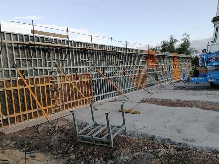 Szare szalunki ustawione pionowo, podparte żółtymi stemplami, widoczny dźwig na budowie w tle.