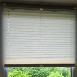 Jasna, plisowana osłona okienna w pozycji częściowo zasłoniętej, przez okno widok na bujną zieleń ogrodu.