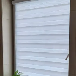 Okno w łazience z zamontowaną białą roletą typu dzień i noc, na parapecie doniczka z paprotką i odświeżacz powietrza.