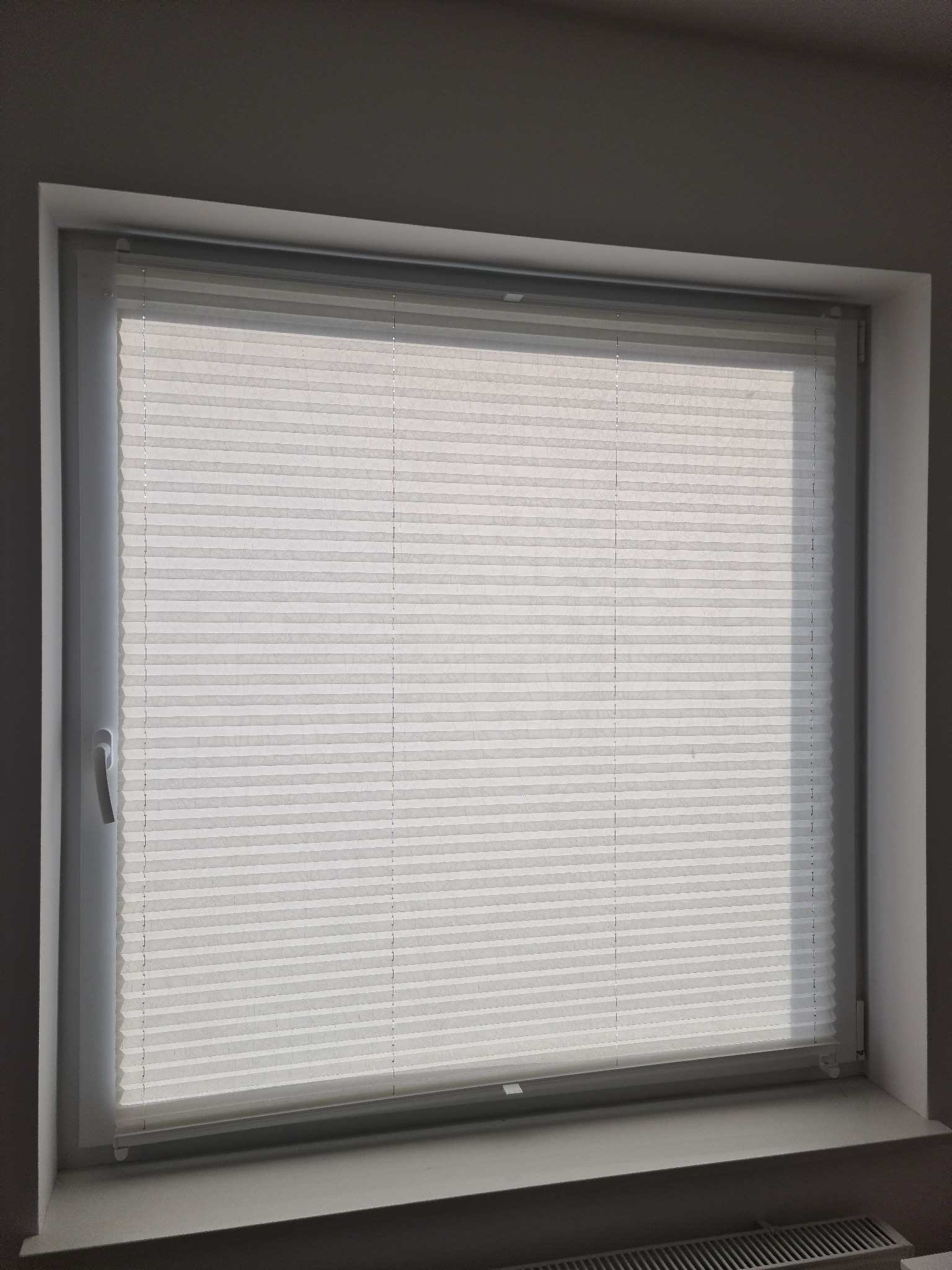 Białe, plisowane osłony okienne zamontowane w oknie PCV, widok z wnętrza pomieszczenia, delikatne światło dzienne.