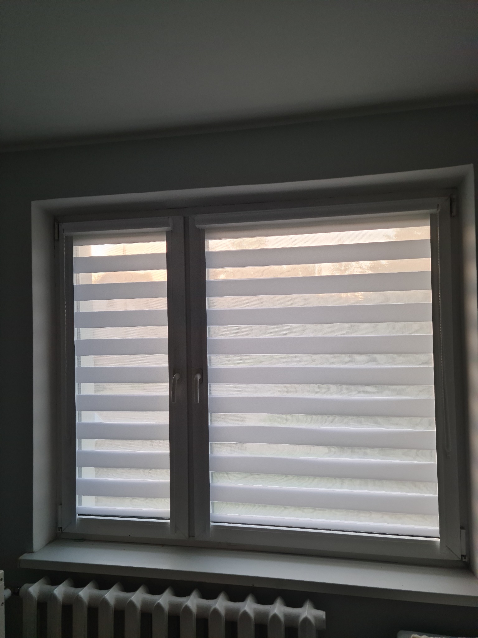 Okno z zamontowanymi białymi roletami z pasami, widoczny grzejnik pod parapetem.