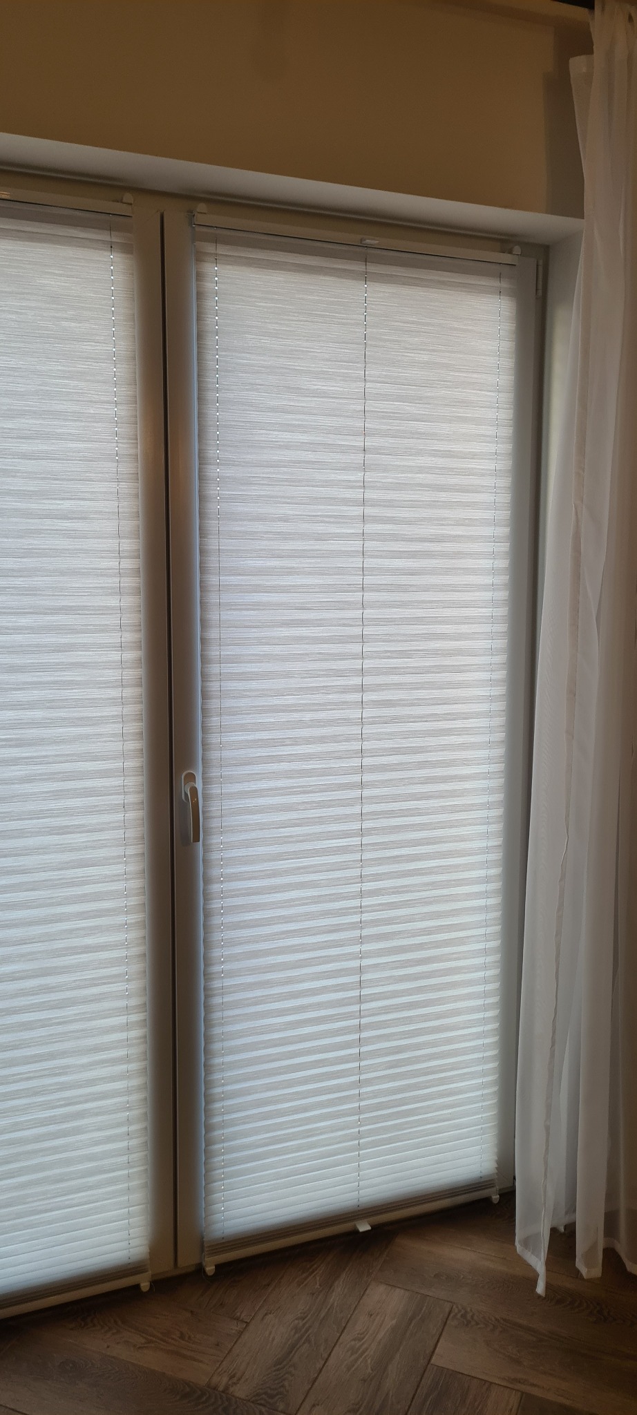 Zamknięte, białe plisy okienne na dwuskrzydłowym oknie, z widoczną klamką i fragmentem firany po prawej stronie.