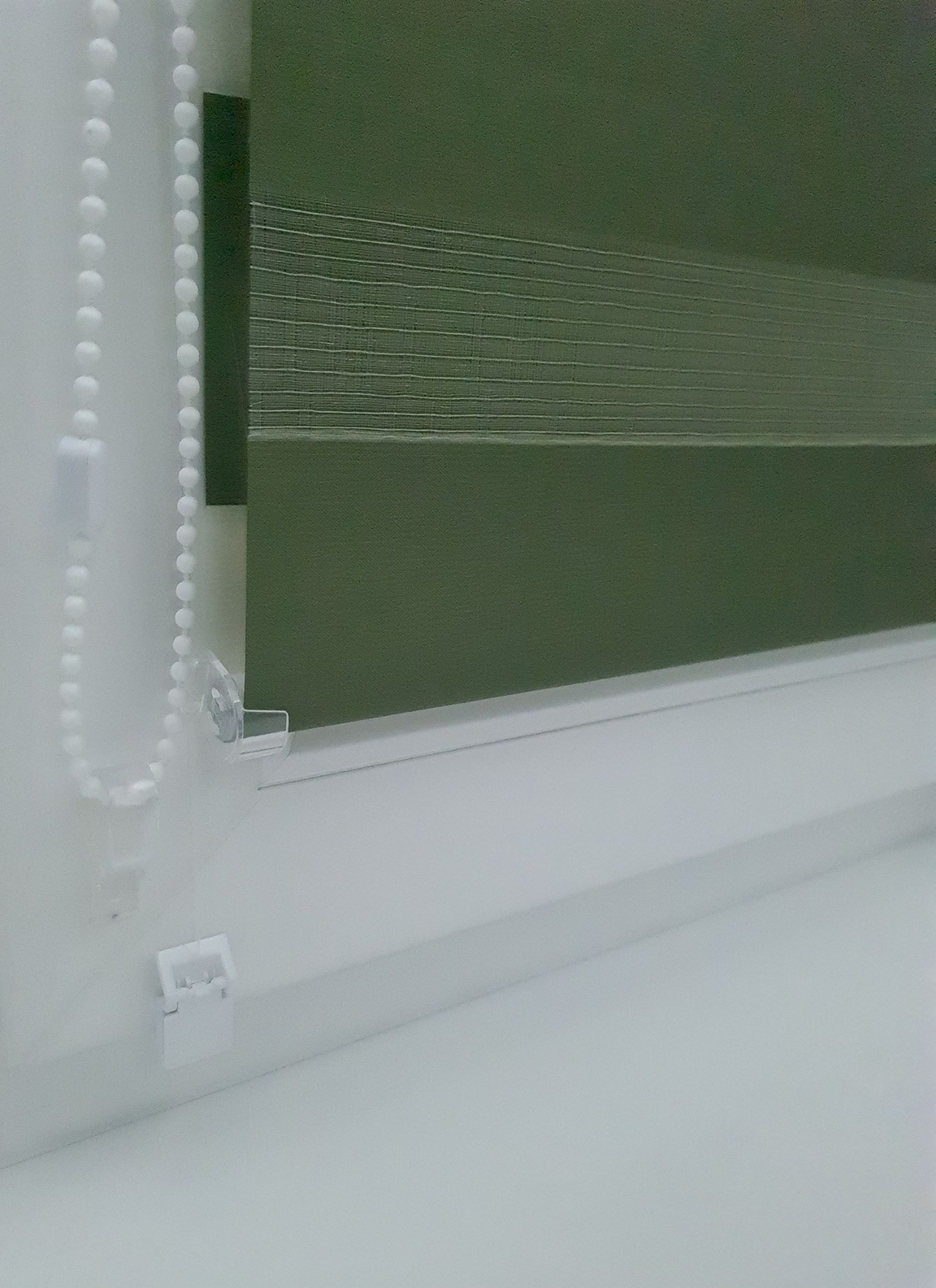 Zaciemniona roleta dzień noc w kolorze oliwkowym, widoczny mechanizm łańcuszkowy i biała rama okienna.