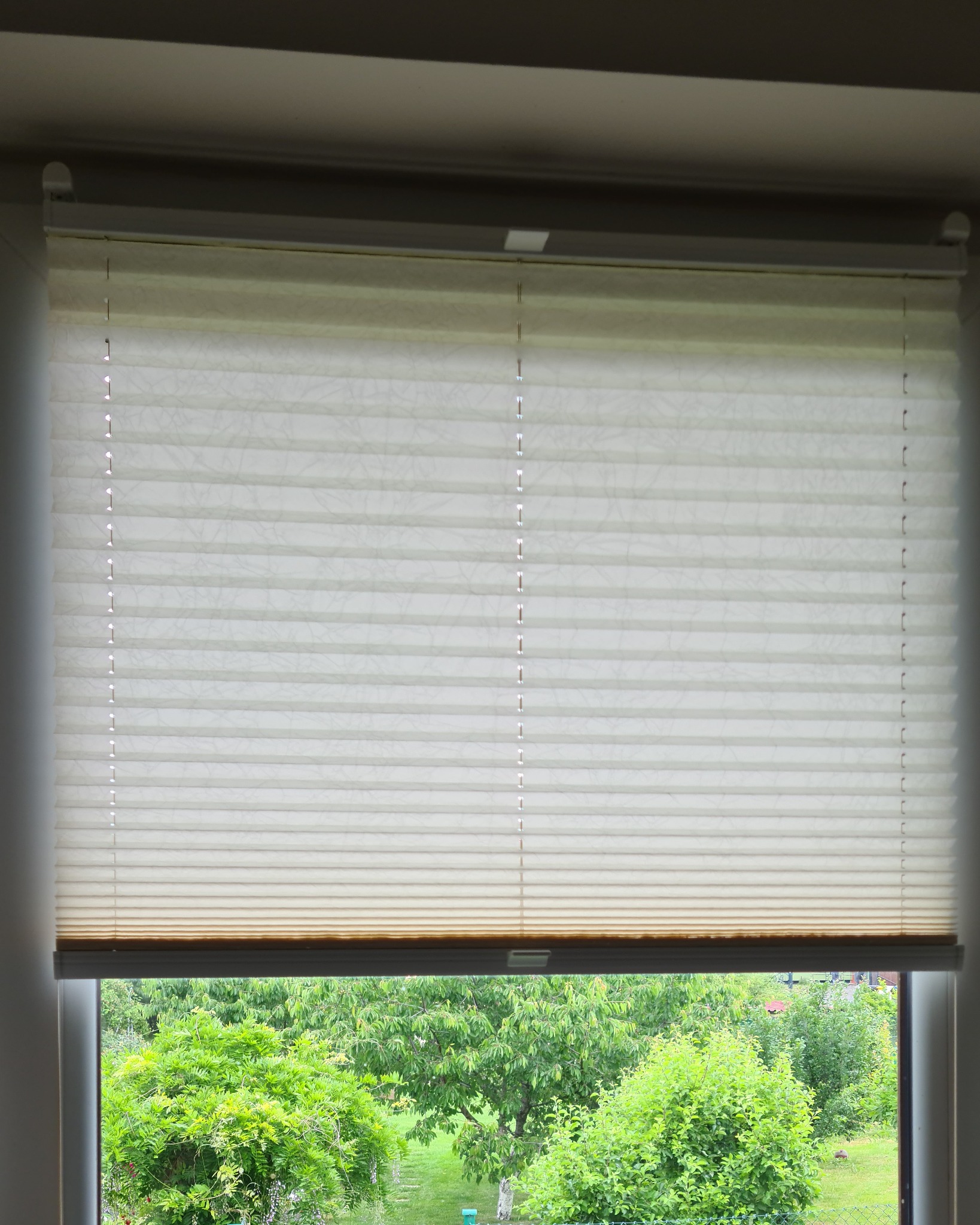 Jasna, plisowana osłona okienna w pozycji częściowo zasłoniętej, przez okno widok na bujną zieleń ogrodu.