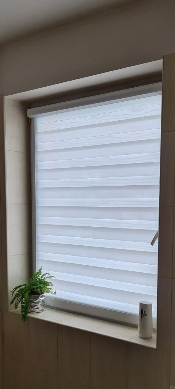 Okno w łazience z zamontowaną białą roletą typu dzień i noc, na parapecie doniczka z paprotką i odświeżacz powietrza.