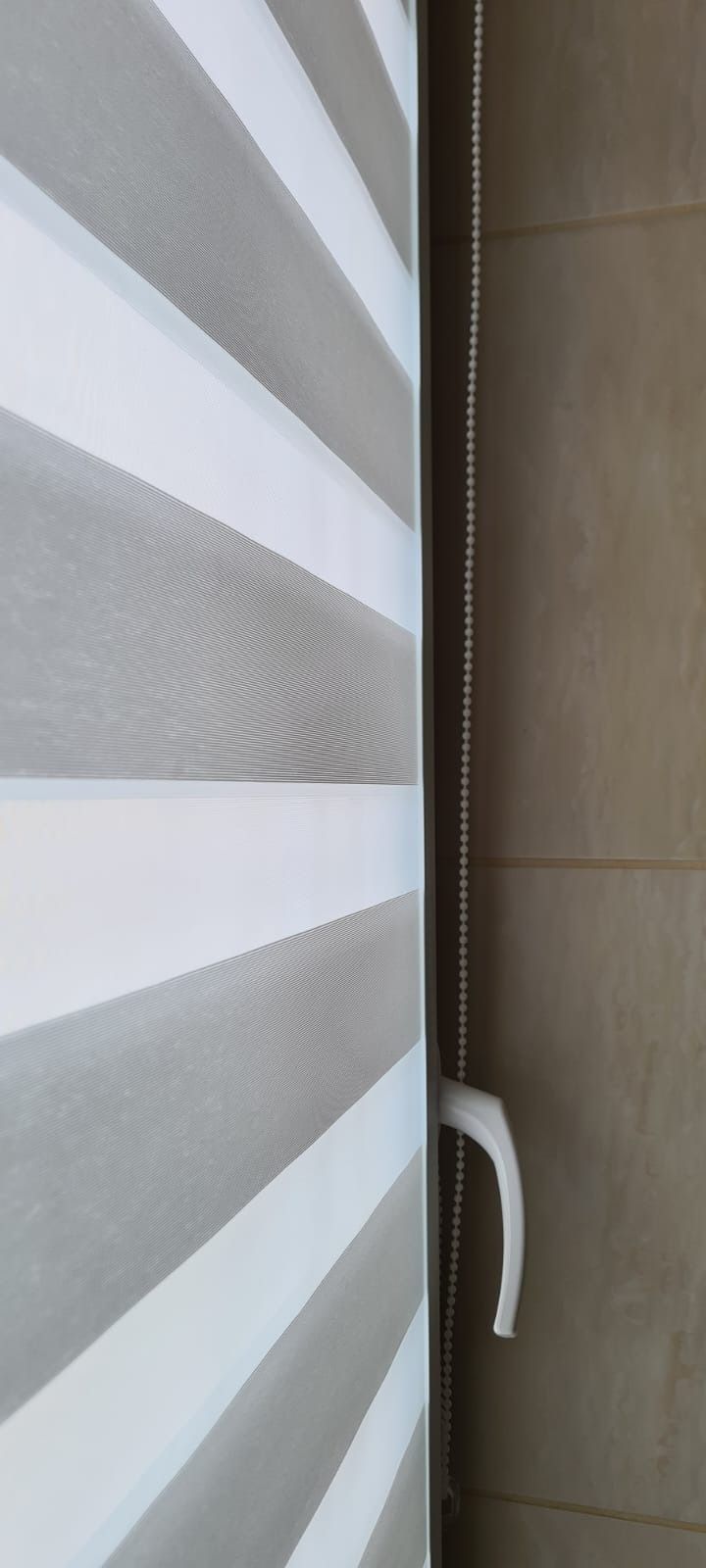 Fragment okna z zamontowaną szarobiałą przesłoną z poziomymi pasami, widoczny mechanizm łańcuszkowy z białym uchwytem.