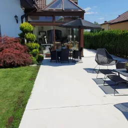 Jasna, gładka posadzka tarasu przylegającego do domu z oranżerią, z czarnymi meblami ogrodowymi i parasolem.