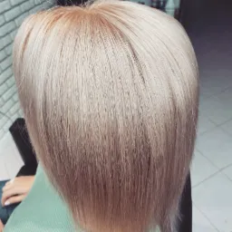 Blond włosy po koloryzacji, widoczne od tyłu, z delikatnym odrostem w odcieniu naturalnego blondu i chłodnymi tonami platyny na końcach, na tle ściany z białej cegły.