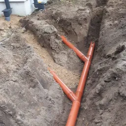 Pomarańczowe rury drenażowe w wykopie ziemnym, widoczne przyłącza, obok ściana budynku i narzędzia.