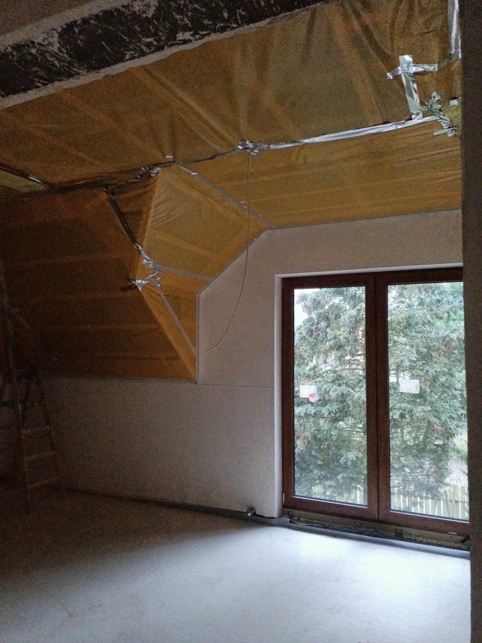 Poddasze w trakcie ocieplania, widoczne żółte płyty izolacyjne przymocowane do skośnego dachu za pomocą srebrnej taśmy, surowa ściana i okno z widokiem na drzewo.