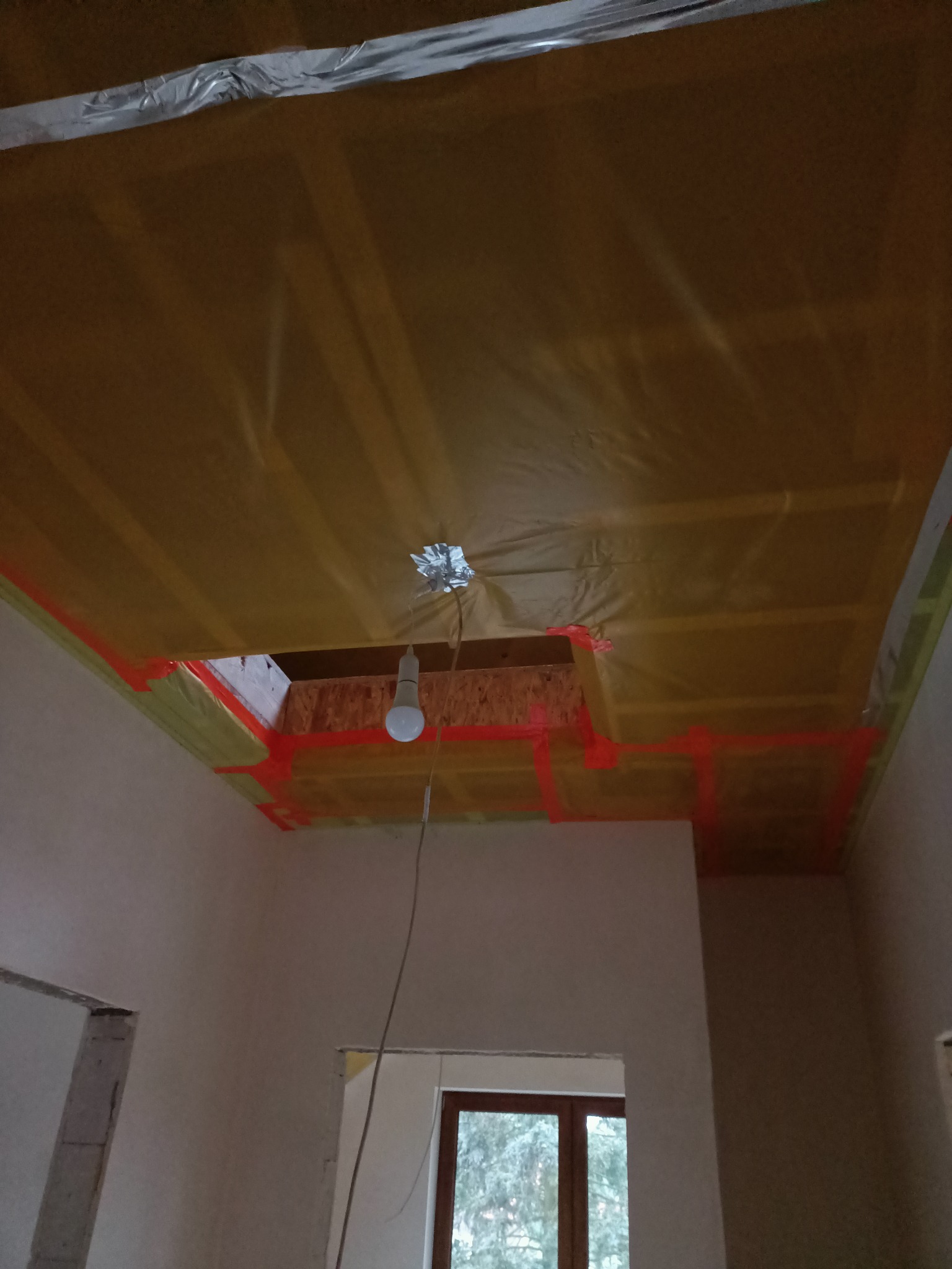 Poddasze w trakcie ocieplania, widoczne żółte folie i drewniane belki, zabezpieczone czerwoną taśmą. W suficie otwór z żarówką. Prace wykończeniowe w pomieszczeniu.