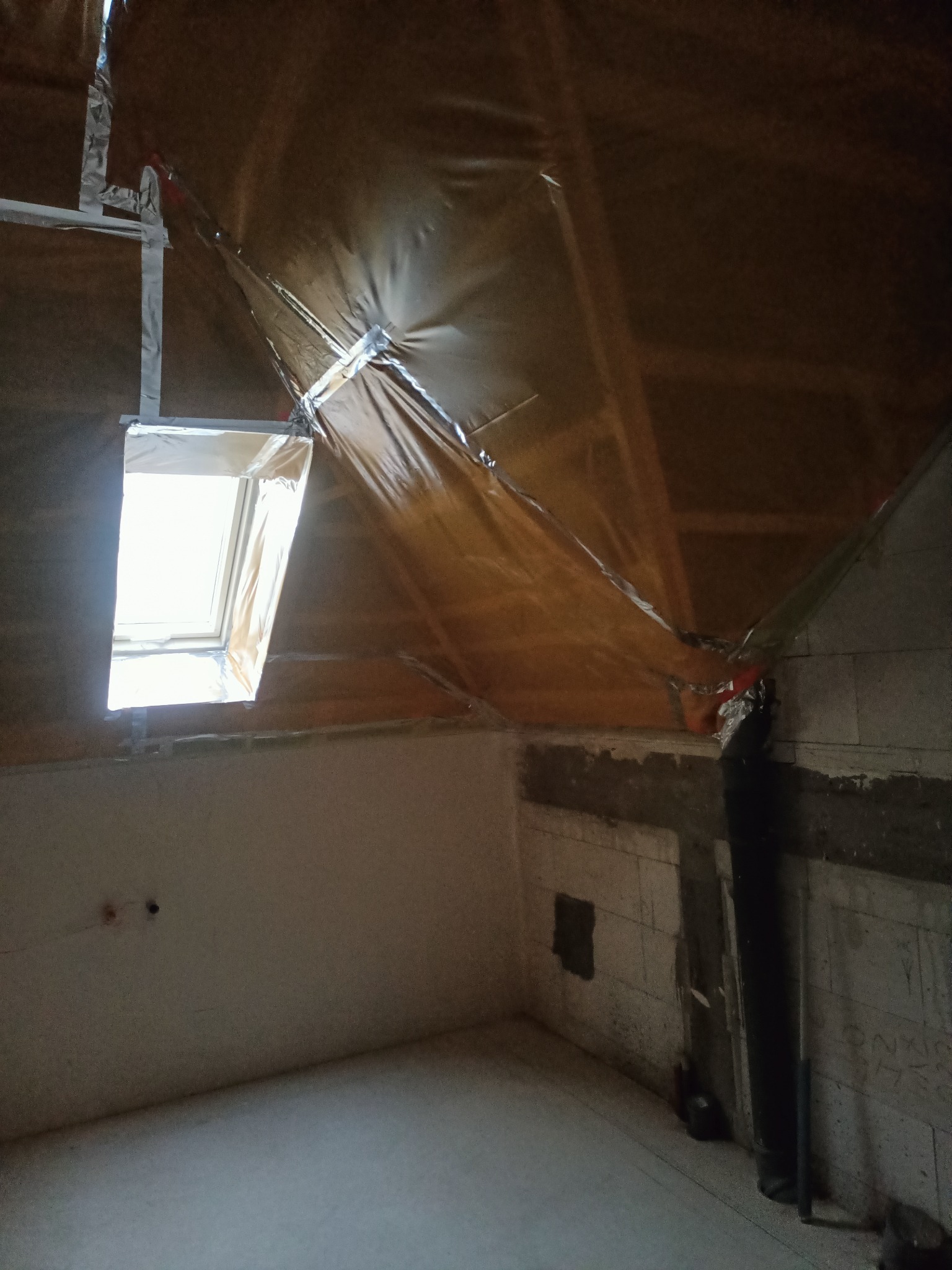 Poddasze w trakcie ocieplania folią paroizolacyjną, widoczne okno dachowe, surowa ściana z betonu komórkowego z instalacją elektryczną w tle.