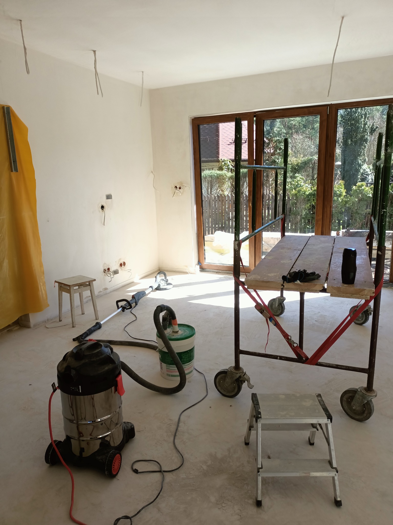 Pokój w trakcie prac wykończeniowych: szpachlowanie i szlifowanie ścian, widoczne rusztowanie, szlifierka do gładzi, odkurzacz przemysłowy, ślady pyłu na podłodze, okno balkonowe z widokiem na ogród.