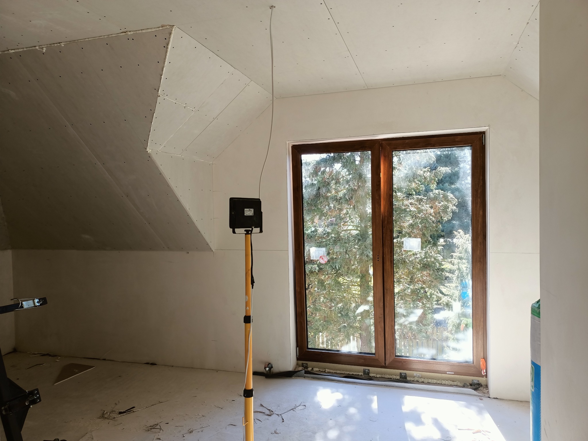 Poddasze po montażu płyt gipsowo-kartonowych, widoczne szpachlowane łączenia, drewniane okno z widokiem na drzewa, lampa budowlana na statywie.