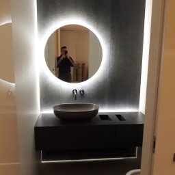Vadym Rusetskyi - Nowoczesna łazienka z okrągłym lustrem z podświetleniem LED, ciemnoszara ściana z efektem betonu, misa umywalkowa w kolorze taupe i ciemna szafka pod umywalką z podświetleniem od dołu. W lustrze...