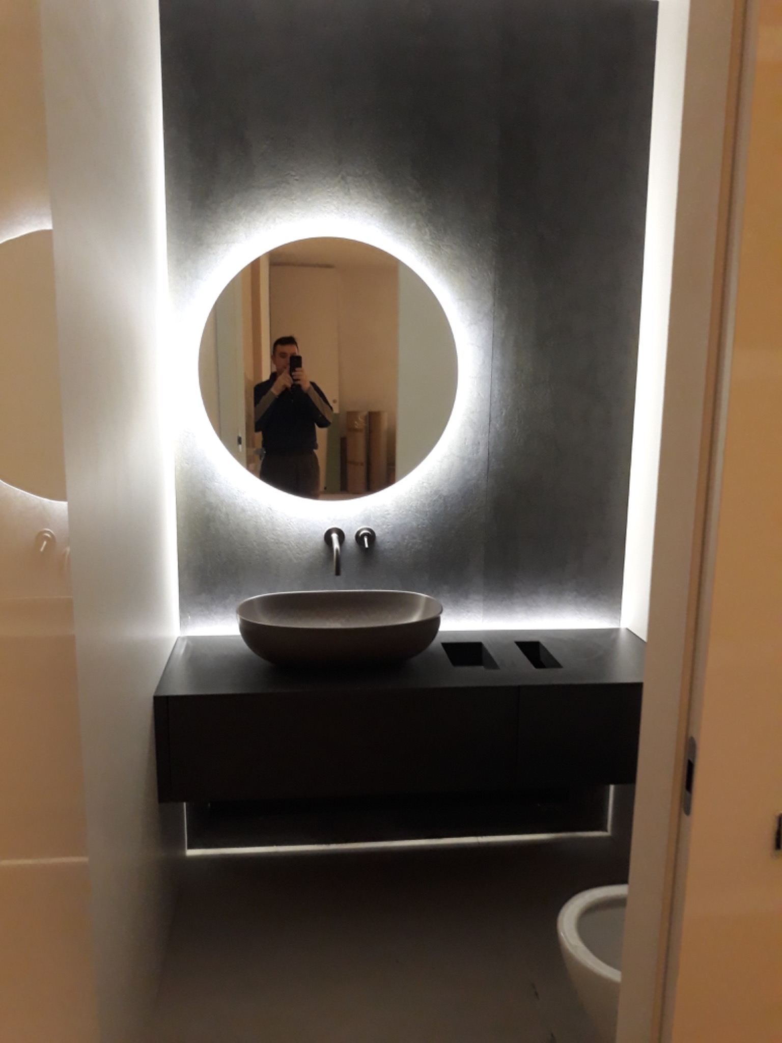 Nowoczesna łazienka z okrągłym lustrem z podświetleniem LED, ciemnoszara ściana z efektem betonu, misa umywalkowa w kolorze taupe i ciemna szafka pod umywalką z podświetleniem od dołu. W lustrze...