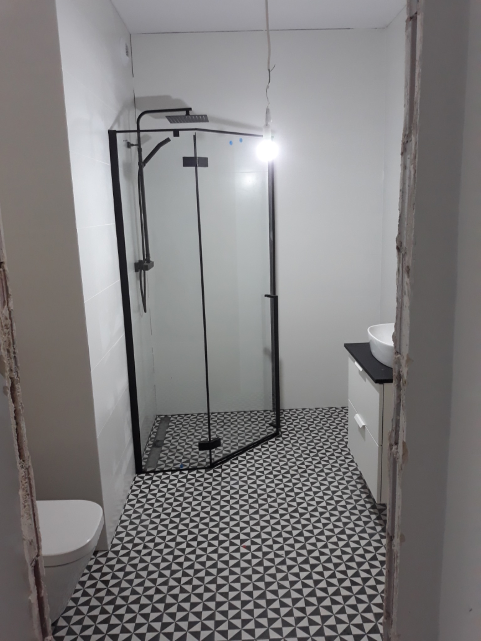 Nowoczesna łazienka z czarną kabiną prysznicową typu walk-in, białą umywalką nablatową i geometryczną podłogą w czarno-białe trójkąty, widoczny surowy fragment ściany przed wykończeniem.