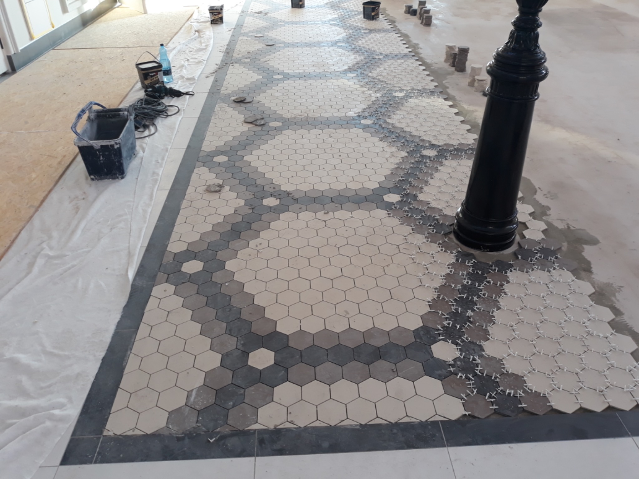 Układanie mozaiki heksagonalnej w odcieniach szarości i beżu na podłodze, widoczne krzyżyki dystansowe, kubeł z zaprawą i kable elektryczne w tle.