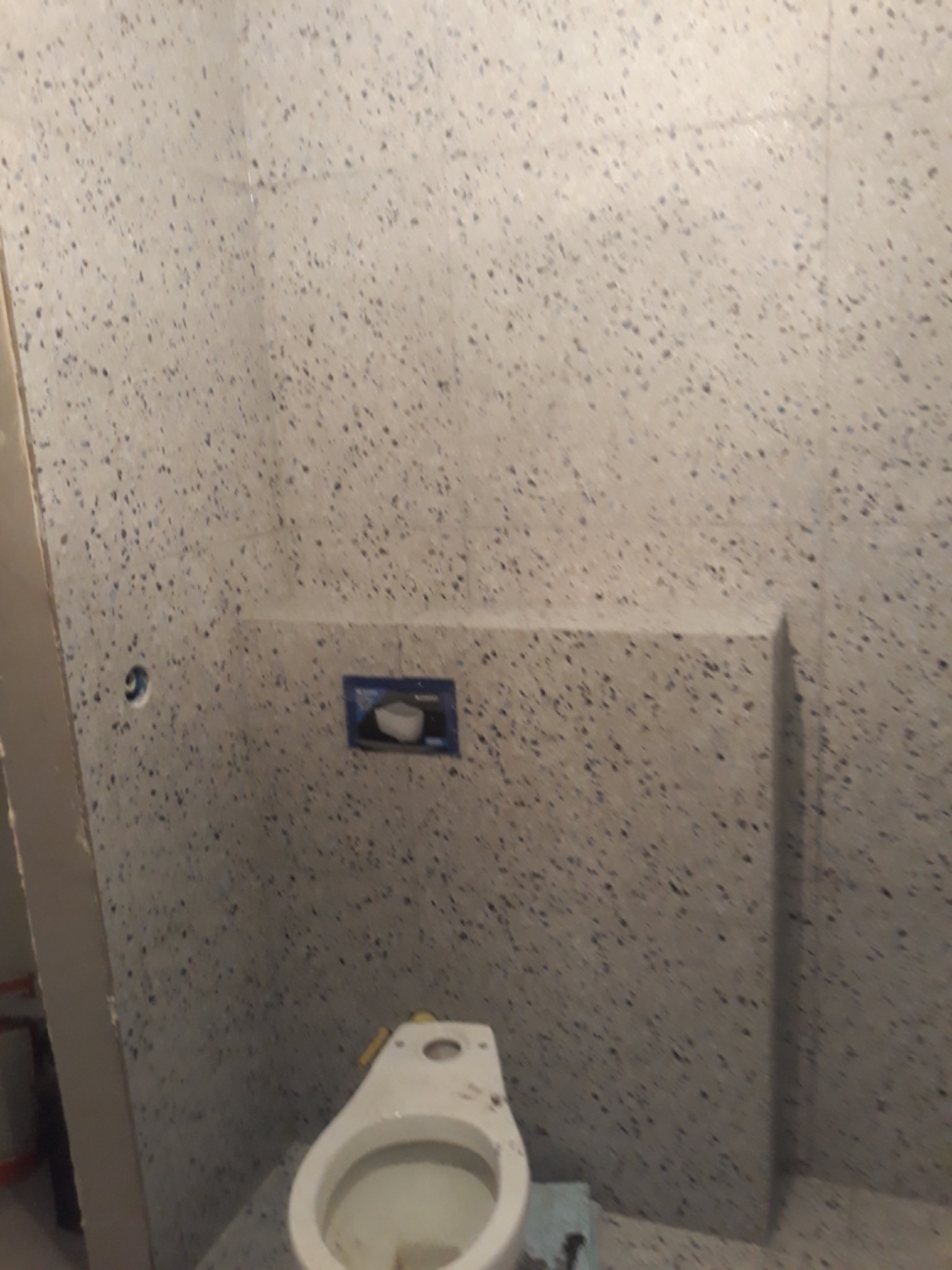 Łazienka w trakcie wykańczania: zamontowany stelaż podtynkowy WC z widocznym otworem na przycisk spłukujący i częściowo ułożone płytki imitujące lastryko na ścianach.